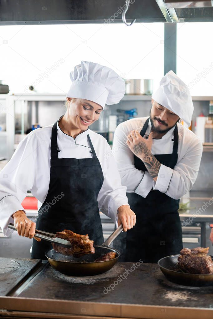Asian chef standing near colleague cooking meat on cooktop in kitchen 