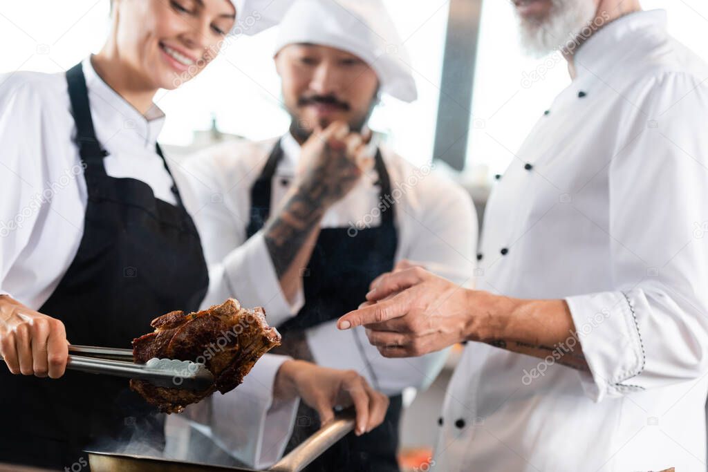 Chef pointing at roasted meat near interracial colleagues in kitchen 