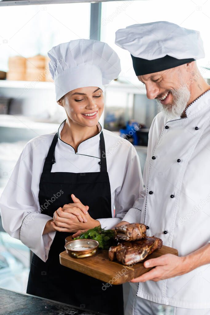 Smiling chef holding roasted meat on cutting board near colleague in apron 