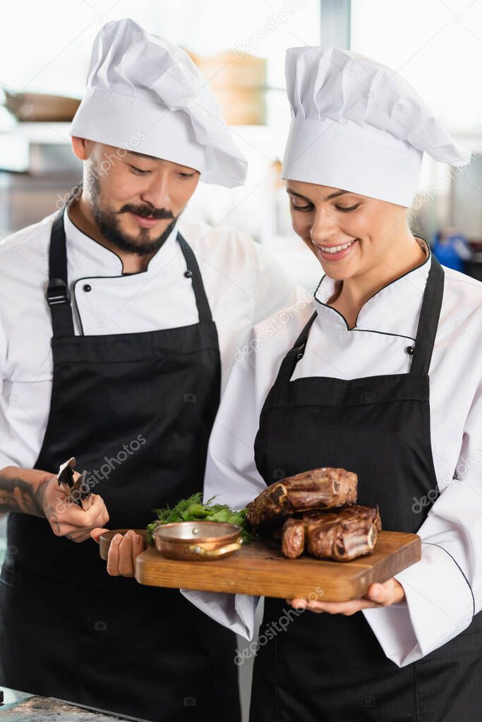 Asian chef holding tongs near cheerful colleague with roasted meat on cutting board 