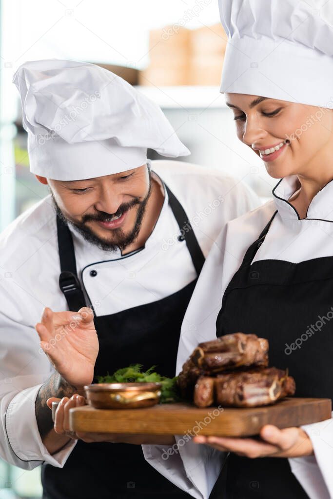 Smiling asian chef showing okay gesture near colleague with roasted meat 