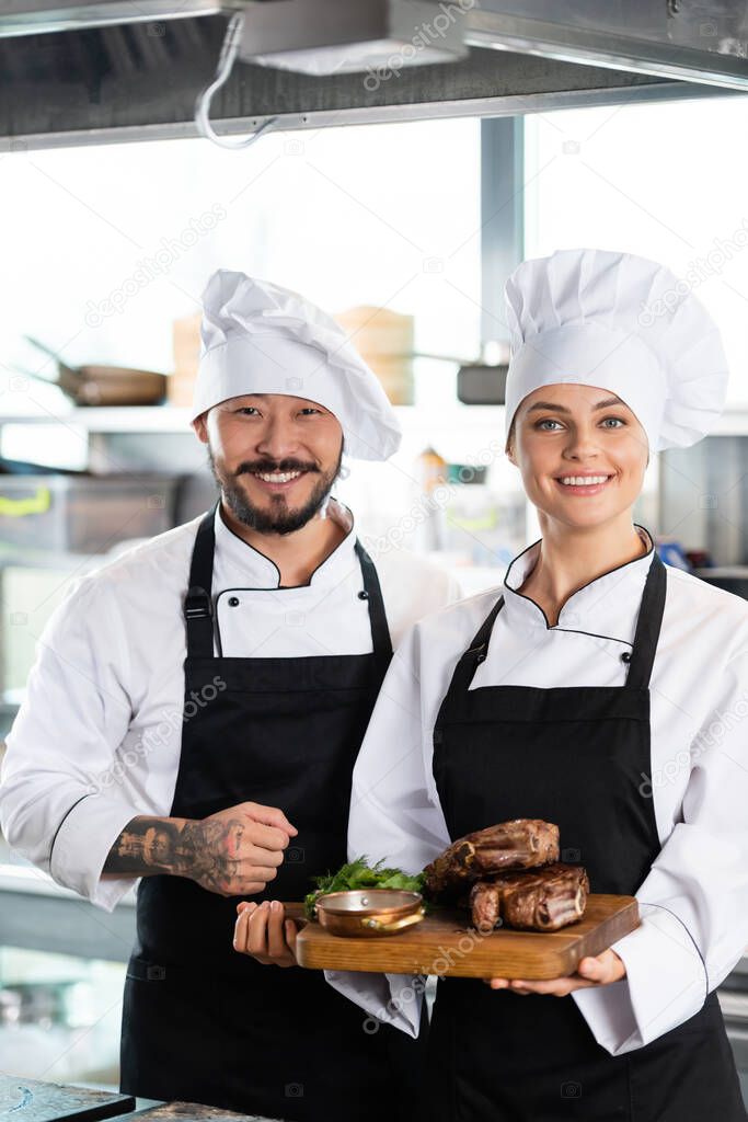 Tattooed asian chef standing near colleague with roasted meat on cutting board in kitchen 
