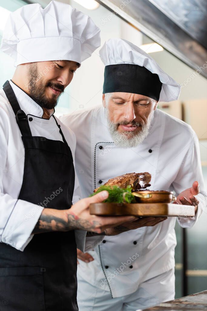 Mature chef smelling roasted meat near asian colleague in kitchen 