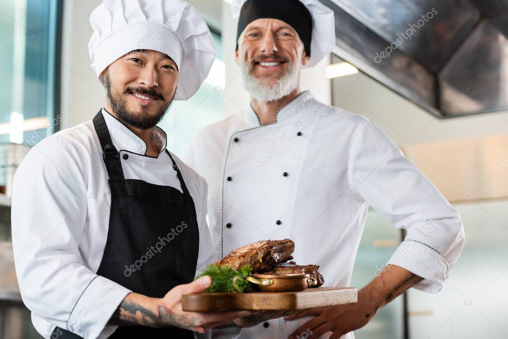 Smiling asian chef holding cutting board with roasted meat near colleague in kitchen 