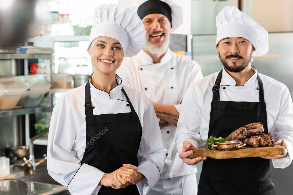 Asian chef holding roasted meat near cheerful colleagues in kitchen 