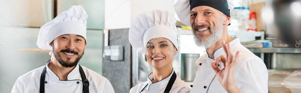 Smiling chef showing okay gesture near interracial colleagues, banner 