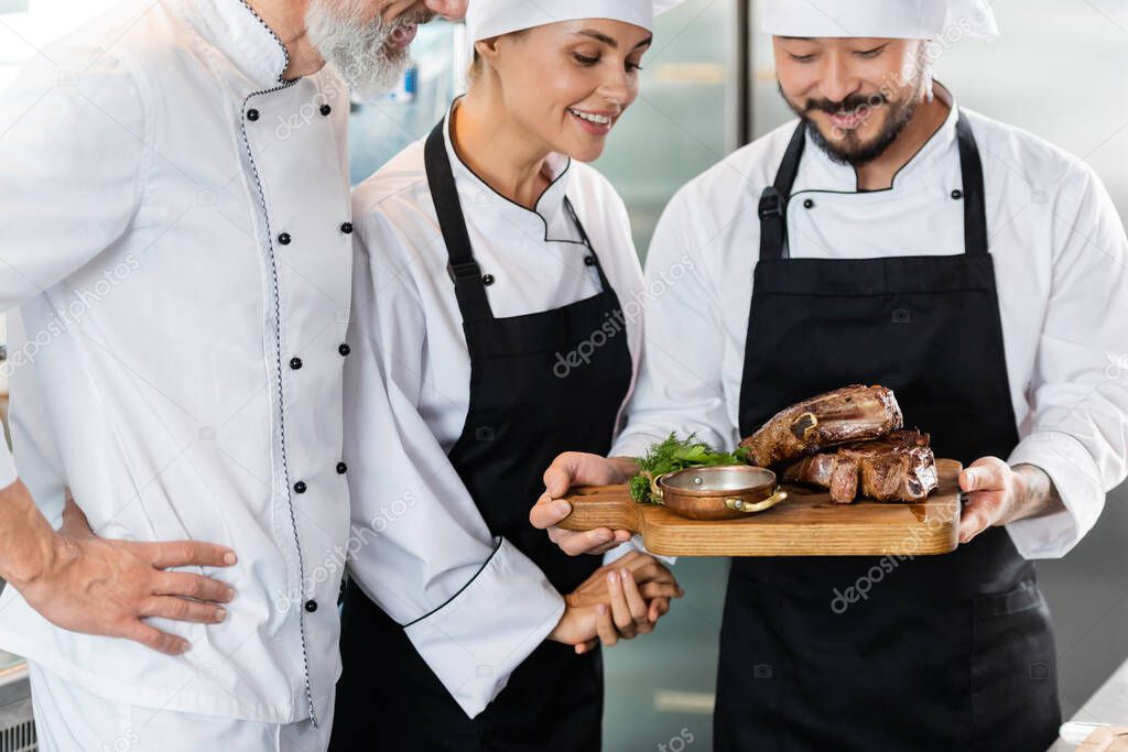 Smiling interracial chefs looking at tasty meat on cutting board in kitchen 
