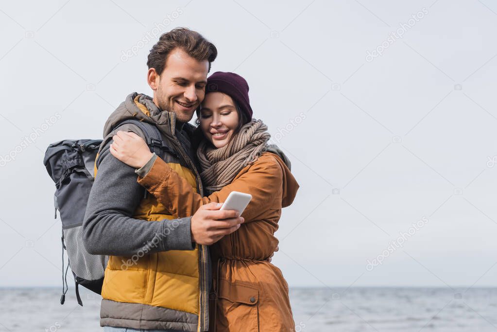 smiling couple of travelers looking at smartphone near sea