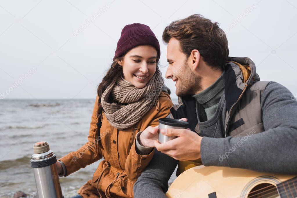 smiling man taking warm drink from woman with thermos while sitting near sea