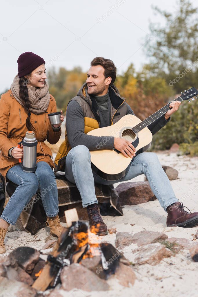 woman with thermos and man with acoustic guitar sitting on stones near bonfire