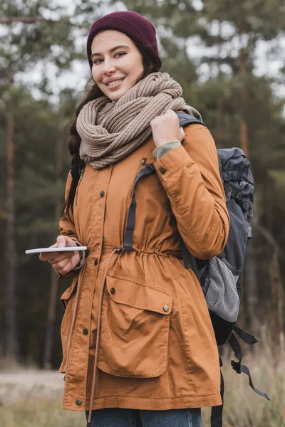 森の中をハイキングしながらカメラを見ながらバックパックと携帯電話を持つ正の若い女性 — ストック写真