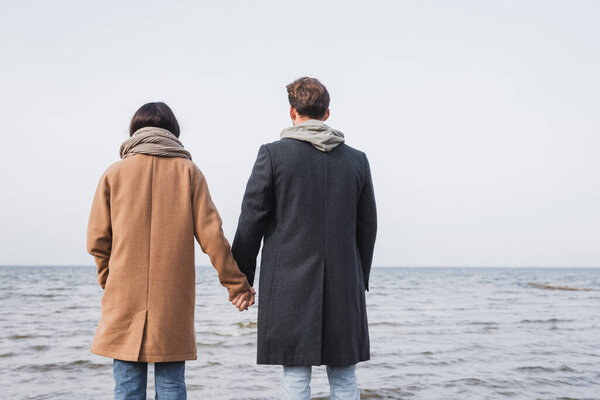 back view of couple in autumn outfit holding hands while standing near sea