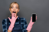 Astonished blonde plumber in rubber gloves holding cellphone with blank screen isolated on grey 