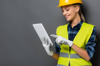 Güvenlik yeleği giymiş gülümseyen sarışın müteahhit ve eldivenler gri tableti işaret ediyor. 