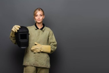Young blonde welder in gloves holding mask and looking at camera on grey background clipart