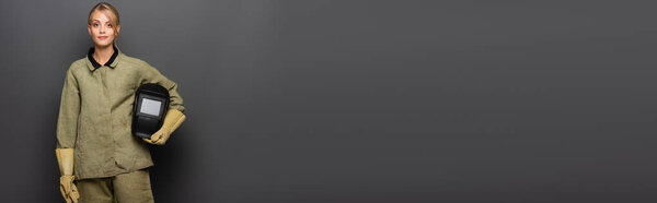 Young blonde welder in uniform holding helmet and looking at camera on grey background, banner 