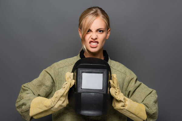 Angry blonde welder in gloves and uniform holding helmet isolated on grey 
