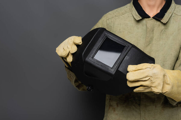 Cropped view of welder in uniform and gloves holding mask on grey background