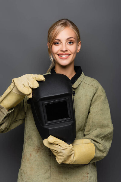 Happy blonde welder in gloves holding helmet and looking at camera isolated on grey 