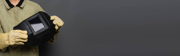 Cropped view of welder in gloves holding helmet with visor on grey background, banner 