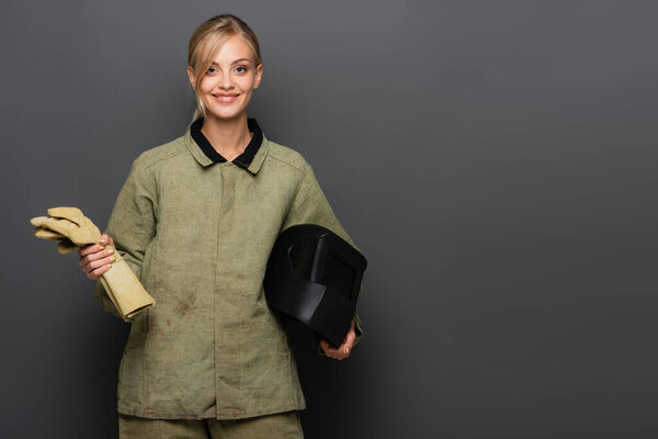 Cheerful blonde welder holding gloves and protective helmet on grey background