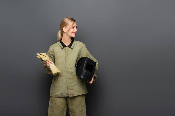 Young blonde welder holding helmet and gloves while looking away on grey background