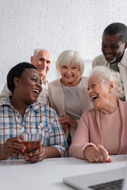 Huzurevinde gülümseyen arkadaşlarının ve laptopun yanında çay tutan Afrikalı Amerikalı kadın. 