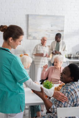 Gülümseyen hemşire huzurevindeki yaşlı Afrikalı Amerikalı kadına dokunuyor. 