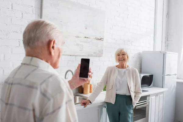 Mujer Mayor Sonriente Mirando Amigo Borroso Con Teléfono Inteligente Hogar —  Fotos de Stock