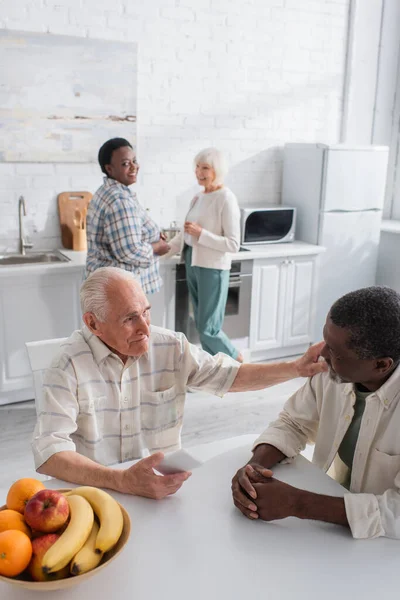 Interracial senior men with smartphone talking near fruits and blurred women in nursing home
