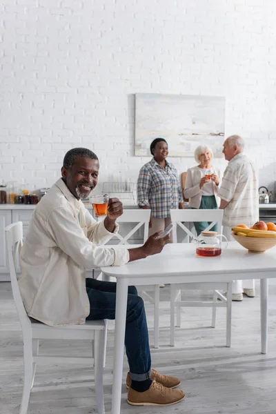 笑顔アフリカ系アメリカ人男性ホールディングお茶とスマートフォン近くぼやけた友人で老人ホーム — ストック写真