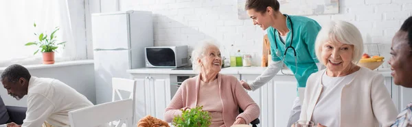 老人ホームの異人種間の年金受給者の近くの車椅子の高齢女性と話す看護師 バナー — ストック写真