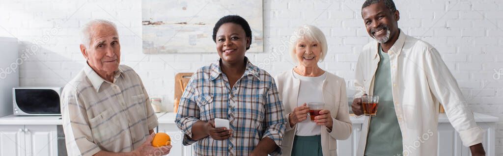 Smiling interracial senior people with tea and smartphone in nursing home, banner 