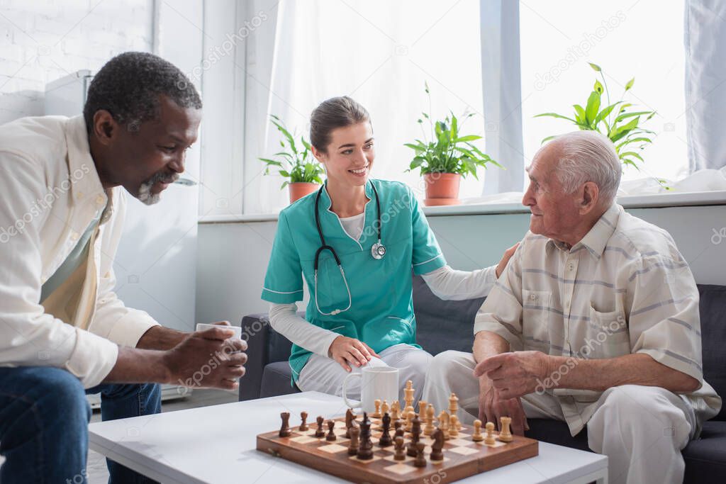 Smiling nurse sitting near interracial patients and chess in nursing home 