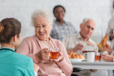 Huzurevinde bulanık hemşire ile konuşurken elinde çay tutan pozitif yaşlı kadın. 