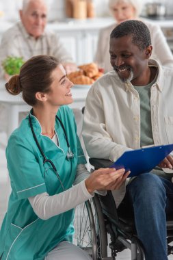 Huzurevinde tekerlekli sandalyedeki Afrikalı Amerikalı bir hastanın yanında pano tutan neşeli hemşire. 