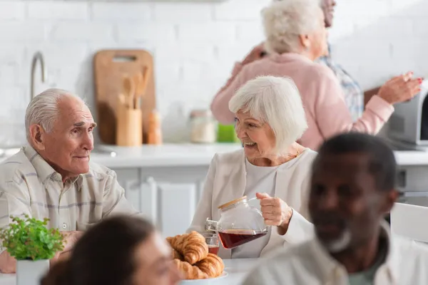Positiv Äldre Kvinna Som Håller Tekanna Nära Vän Och Croissanter — Stockfoto