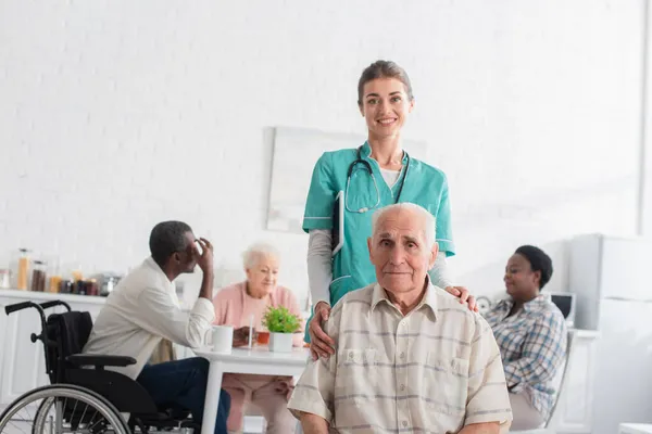 Enfermera Sonriente Mirando Cámara Cerca Pacientes Mayores Personas Interracial Asilo — Foto de Stock