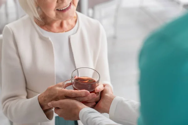 Cropped view of nurse holding hands of smiling patient with tea in nursing home