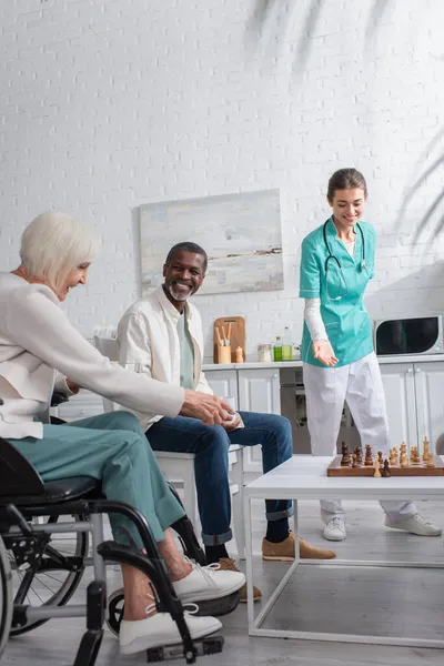 Enfermera Sonriente Señalando Tablero Ajedrez Cerca Pacientes Interracial Asilo Ancianos — Foto de Stock