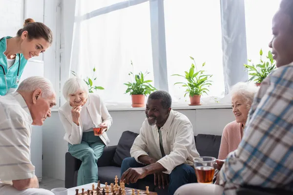 Pacientes Inter Raciais Positivos Com Chá Jogando Xadrez Perto Enfermeira — Fotografia de Stock