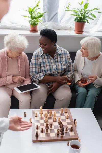 老人ホームでお茶とチェスと異人種間の友人の近くのデジタルタブレットを指しているシニア女性 — ストック写真