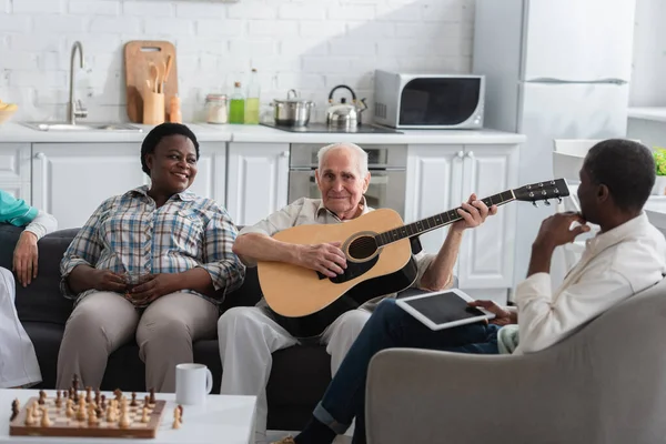 Elderly man playing acoustic guitar near multiethnic friends with tea and digital tablet in nursing home