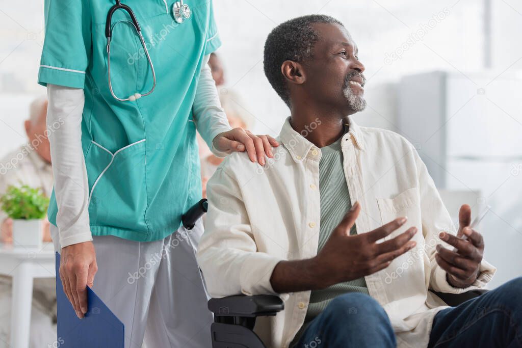 Smiling african american patient holding smartphone while sitting in wheelchair near nurse in nursing home 