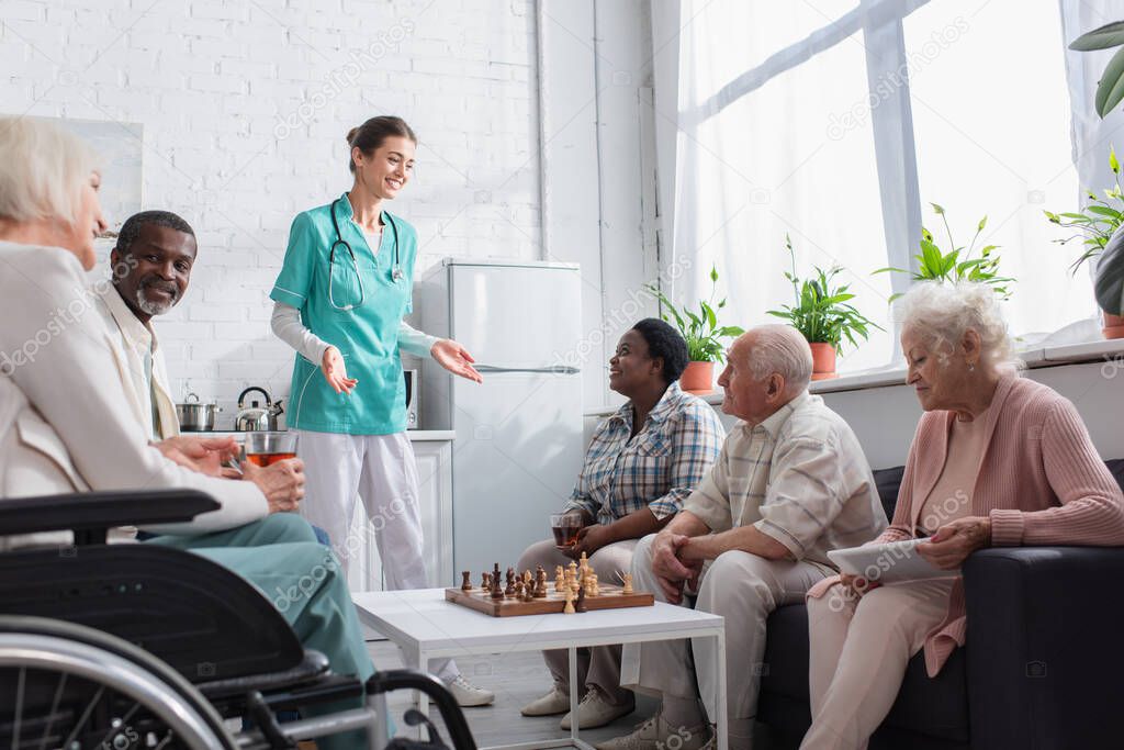 Smiling nurse standing near interracial people with digital tablet and chess in nursing home 