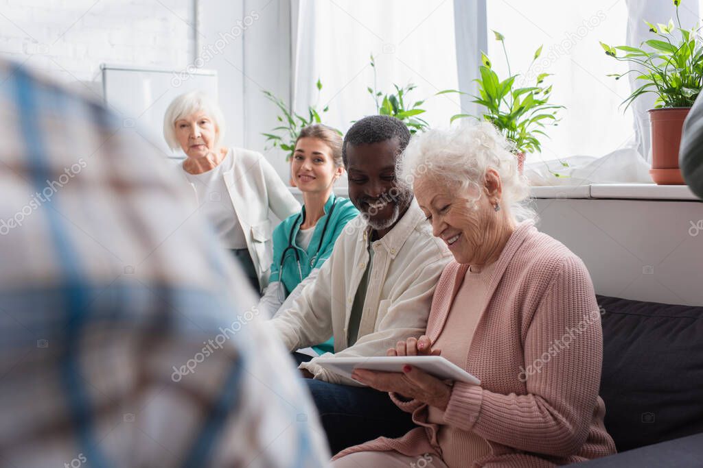Smiling interracial pensioners using digital tablet near nurse in nursing home 