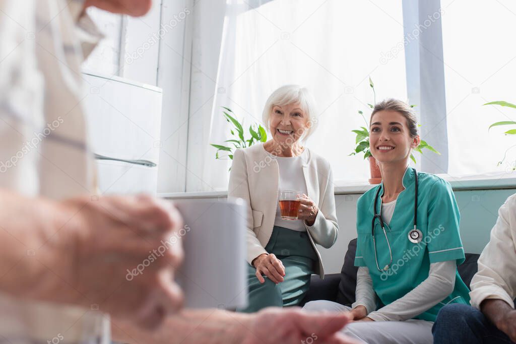 Positive woman with tea and nurse looking at senior man in nursing home 