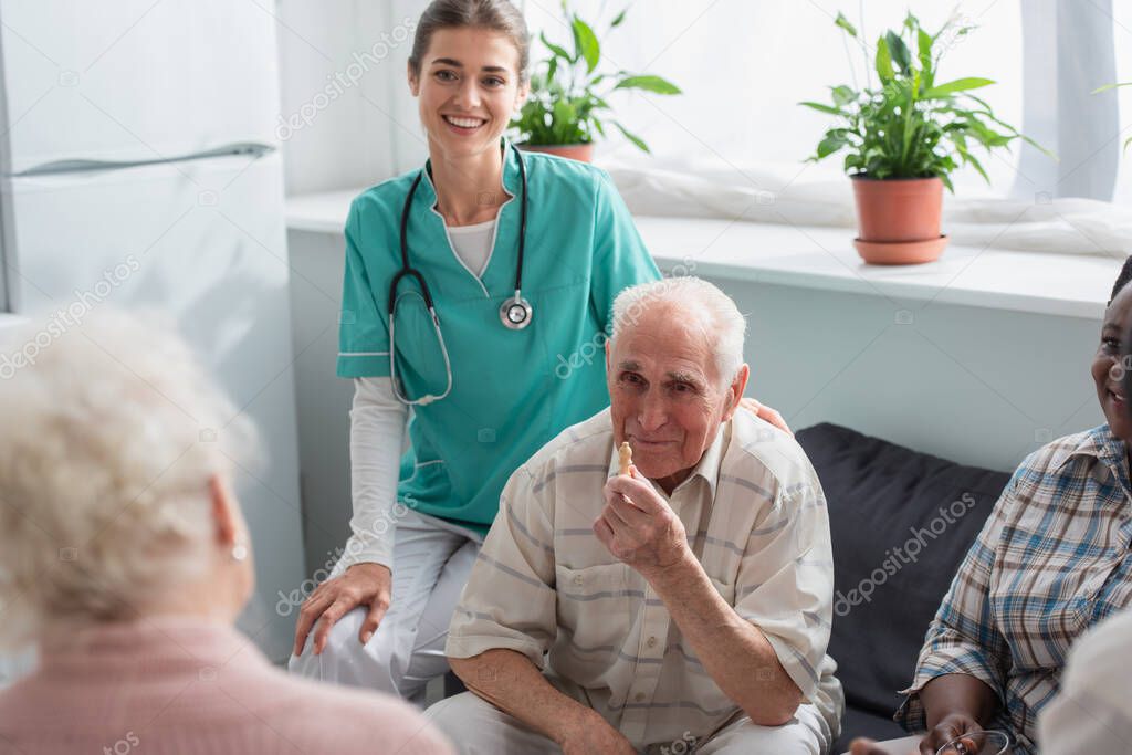 Smiling nurse sitting near senior interracial patients with chess figure in nursing home 