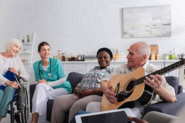 Çok ırklı arkadaşlarının yanında akustik gitar tutan kıdemli bir adam ve huzurevinde hemşire. 