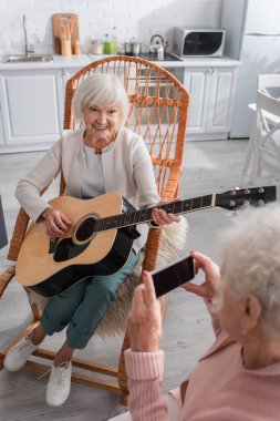 Huzurevinde akıllı telefonlu, akustik gitar çalan gülümseyen yaşlı kadın. 